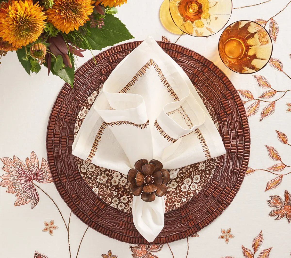 Table setting with a brown woven placemat, a floral-patterned plate, the Kim Seybert Filament Napkin Set of 4 in White & Brown secured with a wooden flower napkin ring, sunflowers in a vase, and two amber-colored drinks on an elegant floral tablecloth.