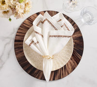 A white and brown embroidered napkin from the Kim Seybert Filament Napkin Set of 4 is elegantly folded and secured with a gold ring atop a beige plate, which rests on a dark wood-grain charger placed on a marble table. These luxurious dining table linens add sophistication to the setting, beautifully complemented by nearby glassware and floral arrangements.