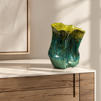 The Sack Vase Green Yellow by David Changar, with its glossy hand-glazed ceramic and organic wavy form, sits on a wooden sideboard. Sunlight casts soft shadows and abstract wall art appears on the left.