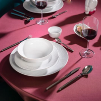 A beautifully arranged dining table adorned with a pink tablecloth showcases the modern elegance of Meissen’s Waves Relief N°41 porcelain dinnerware, complete with white plates and bowls, stylish cutlery, and two glasses of red wine. A small dish of figs and a sophisticated white ceramic accent in the background enhance the refined aesthetic inspired by MEISSEN’s signature design.