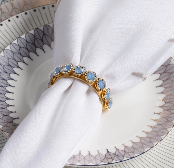 Amaryllis Golden Bar Napkin Ring set with a row of light blue stones and small crystals, holding a white cloth napkin on a patterned plate.