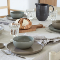 A neatly set dining table showcases Casafina/Costa Nova’s Pacifica Dinnerware in the tranquil Artichoke shade, highlighting elegant gray plates and bowls. The setting is completed with clear glasses, sleek cutlery, a rustic loaf of bread presented on a wooden board, and a dark pitcher—all thoughtfully arranged atop a light tablecloth with folded napkins, creating an understated yet luxurious tablescape.
