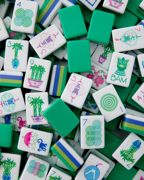 A vibrant collection of Mahjong tiles from the Oh My Mahjong "Mahjong Tiles Set - Jasmine" is displayed in a colorful pile, featuring green-backed tiles along with others adorned with unique designs, numbers, and illustrations like plants, cats, and bamboo. Acrylic tiles with blue, green, and white stripes are also included.