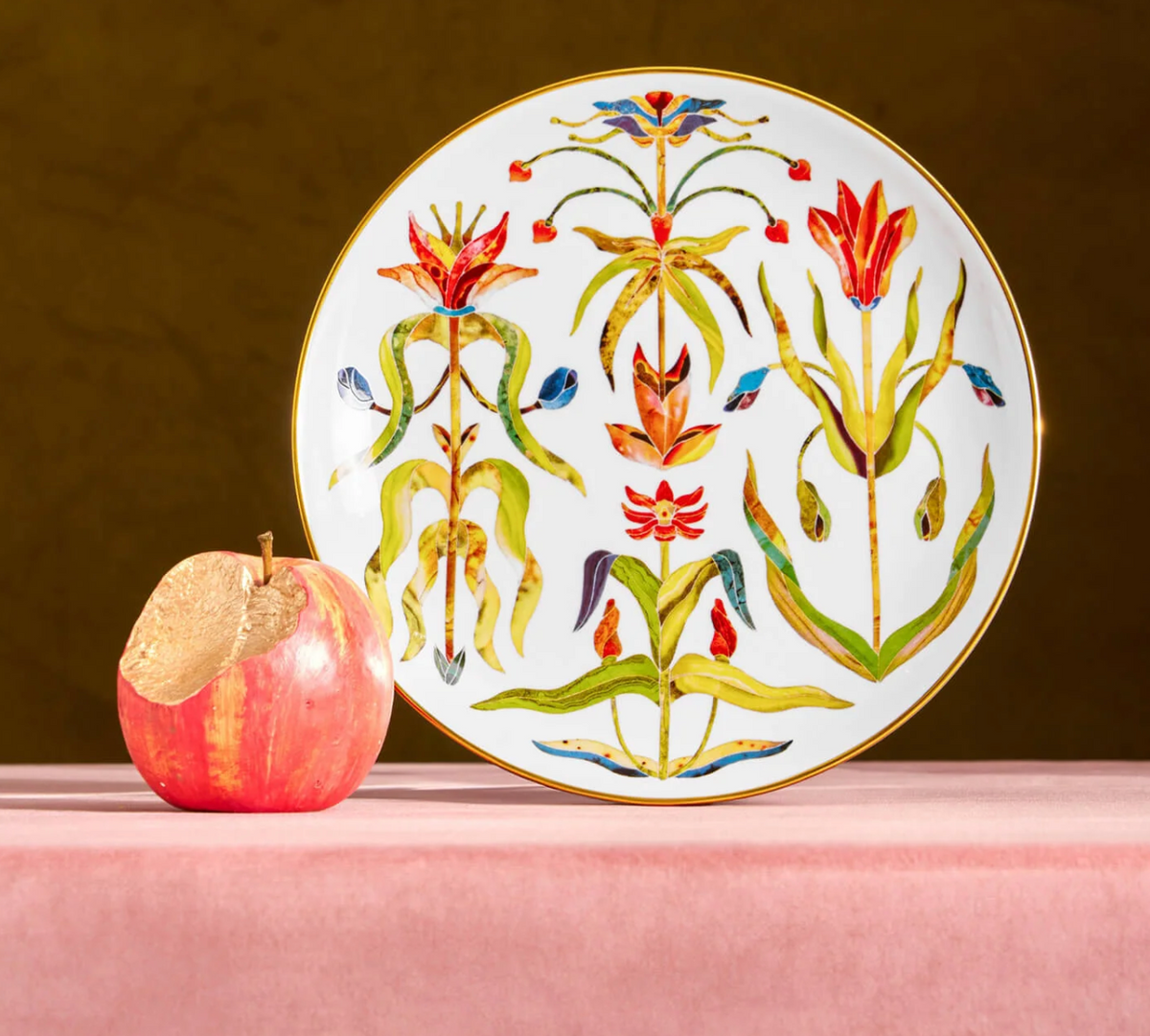 A Grand Tour Dinnerware plate with a 24k gold rim and colorful, stylized floral illustrations, standing next to a red apple with a bite taken out of it.