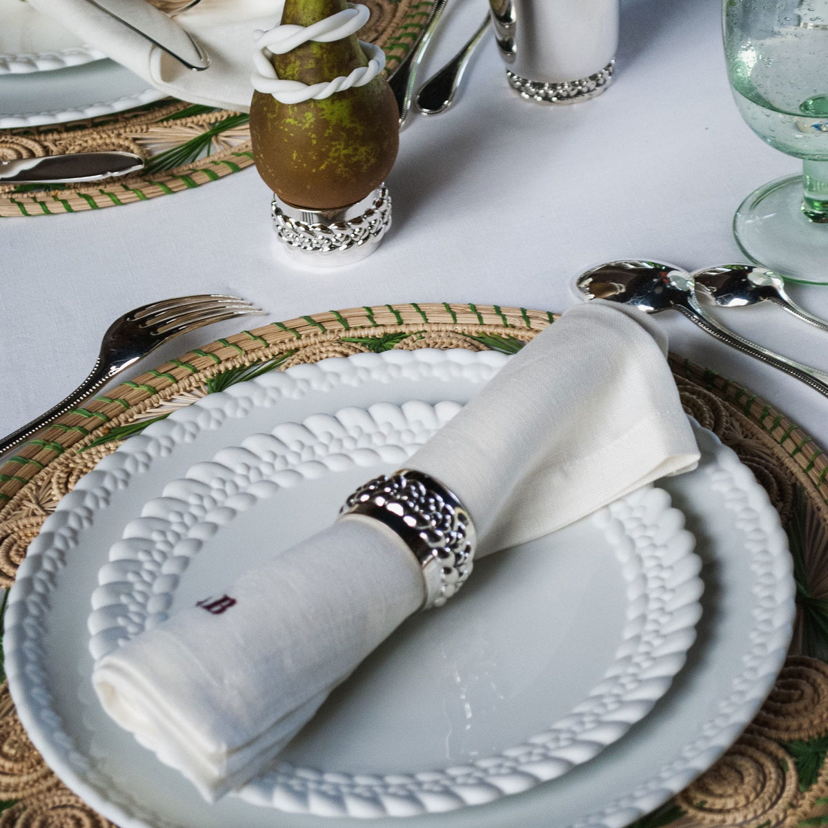 A white napkin rolled through a braided silver-plated Christofle Babylone napkin ring, resting on a white patterned plate atop a woven placemat.