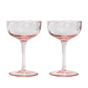 Two clear Puro Cocktail Coupe glasses by Juliska, featuring pink-tinted bases and stems, are placed side by side against a white background—perfect for serving cocktails. This elegant set of two comes beautifully gift boxed, making it an ideal present for any special occasion.