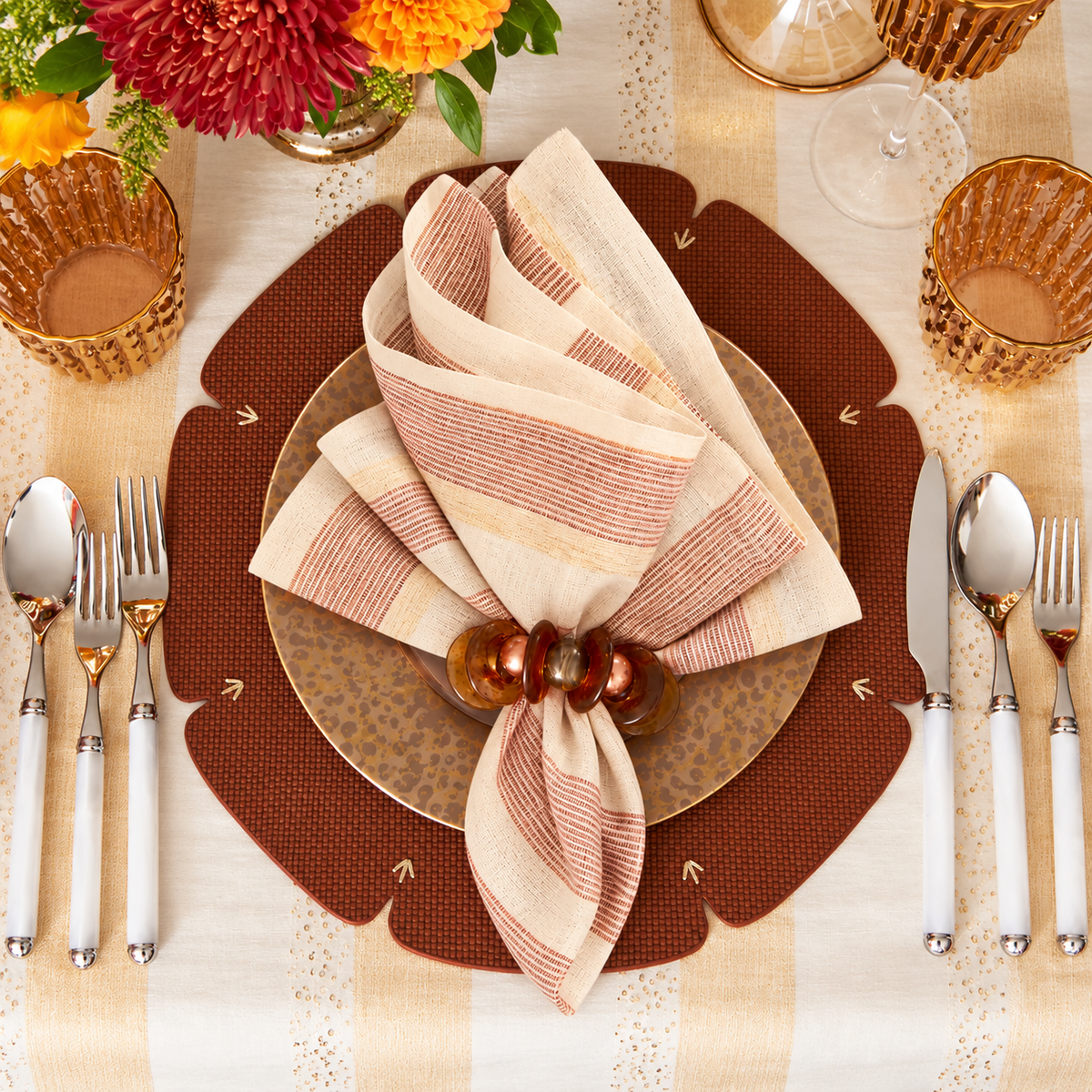 Elevate your table setting with the Kim Seybert Broad Stripe Napkin Set of 4 in Brown & White, elegantly styled in a wooden ring alongside a gold-rimmed plate and gold glassware. Surrounded by silver utensils on a patterned placemat, fresh flowers and golden accents complete the festive look.