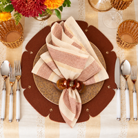 Elevate your table setting with the Kim Seybert Broad Stripe Napkin Set of 4 in Brown & White, elegantly styled in a wooden ring alongside a gold-rimmed plate and gold glassware. Surrounded by silver utensils on a patterned placemat, fresh flowers and golden accents complete the festive look.