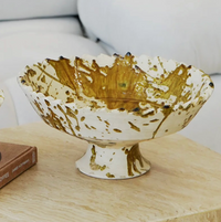 The Jagged Edge Pedestal Fruit Bowl Ochre Splatter by Moroccan White, a handcrafted Moroccan ceramic bowl with a scalloped edge and elegant brown ochre splatter design on a white base, sits on a light wood table near a book and a white couch. Reminiscent of the classic Tamegroute pedestal fruit bowl, this piece combines artisanal charm with contemporary style.