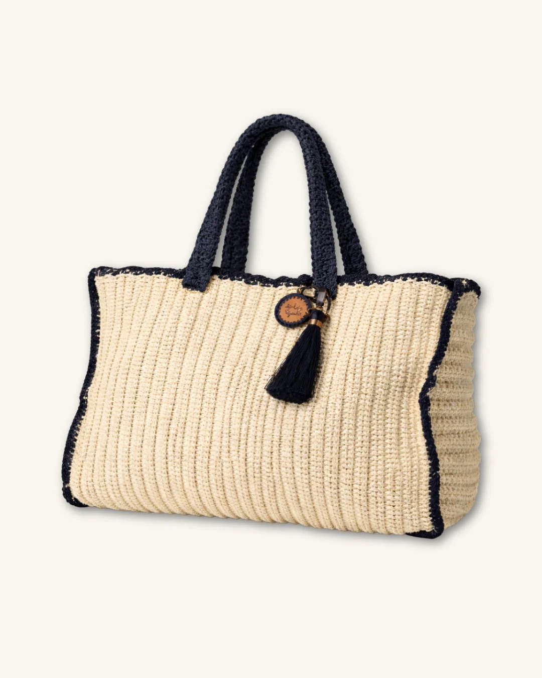 The Arte & Tejido Arte Beach Tote in natural and navy features dark handles, trim, a black tassel, and a round tag—adding subtle bohemian style to your look against a plain light background.