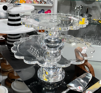 Two Coco Scalloped Cake Servers by Inspired Generations, crafted from resin acrylic, are displayed on a countertop among kitchenware items. Each server features elegant scalloped edges and adds a sophisticated touch to the setting, surrounded by a cake server and various plates and bowls in the background.