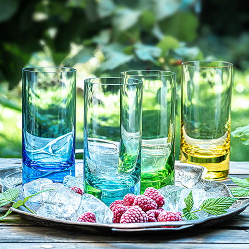 Four colorful Moser Whiskey Spirit Glasses (blue, green, yellow, and aqua) stand on a metal tray with ice cubes and fresh raspberries, set outdoors against lush green foliage.