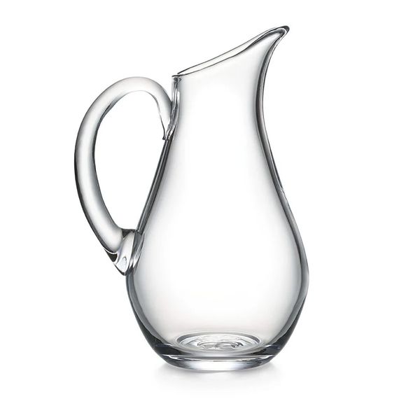 The Simon Pearce Woodstock Pitcher features clear glass, a wide rounded base, curved handle, and narrow angled spout—signature heft from the Woodstock Collection—shown against a white background.