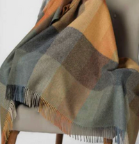 The Sloane Alpaca Throw by Alicia Peru Home, featuring color blocks in gray, blue, brown, and orange with fringed edges, is draped over the back and seat of a light-colored upholstered chair.