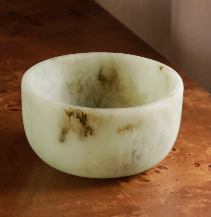 The Beatriz Ball Resin Giada Bowl Jade Swirl - Small features a round silhouette with a light green hue and brown marbling, resting on a wooden surface. Its smooth texture and slightly translucent finish enhance its elegant appearance.