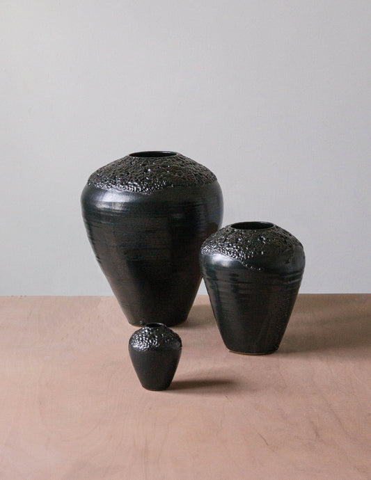 The Esme Crawl Vase - Black Lava by Kalya OD Studio features textured tops in large and medium sizes. Stylish and water safe, these black ceramic vases are arranged on a light wooden surface against a plain background.