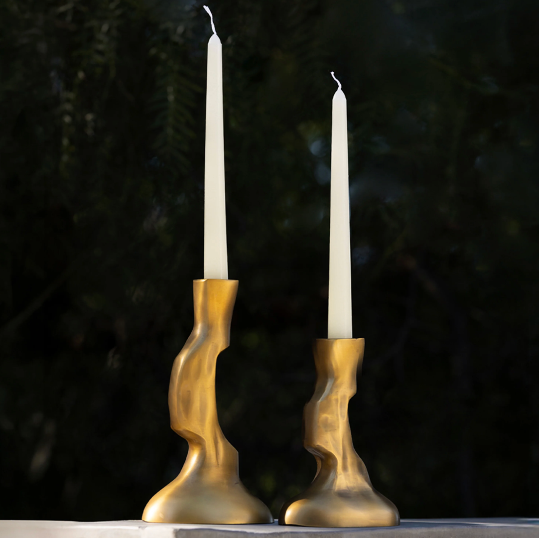 Two white taper candles sit side by side in Frankie Taper Candle Holder Brass - by Dumae, set against a dark, blurred background of foliage.