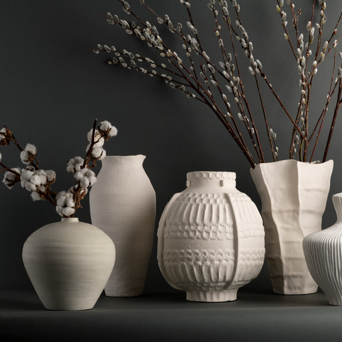 Five off-white Lucille Vases by FlowDecor Inc. of various shapes rest on a dark surface, each showcasing branches with cotton or pussy willow buds against a dark gray background. The artisanal quality and sculptural designs of these vases lend an elegant, handcrafted touch to the serene arrangement.