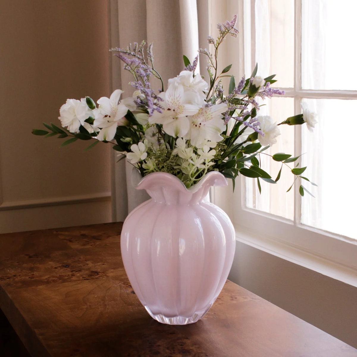 The Beatriz Ball Glass Vento Vase Camellia - Large, filled with white and light purple flowers, sits on a wooden table near a bright window draped with sheer curtains.