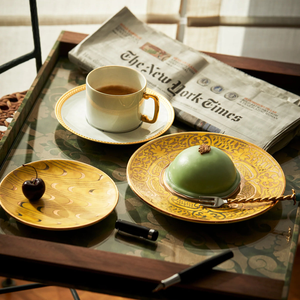 A cup of coffee, a chocolate heart on a L'Objet Fortuny Apollo Canape Plate in yellow, a green dessert under glass, a pen, and The New York Times are stylishly arranged on a tray by the window.