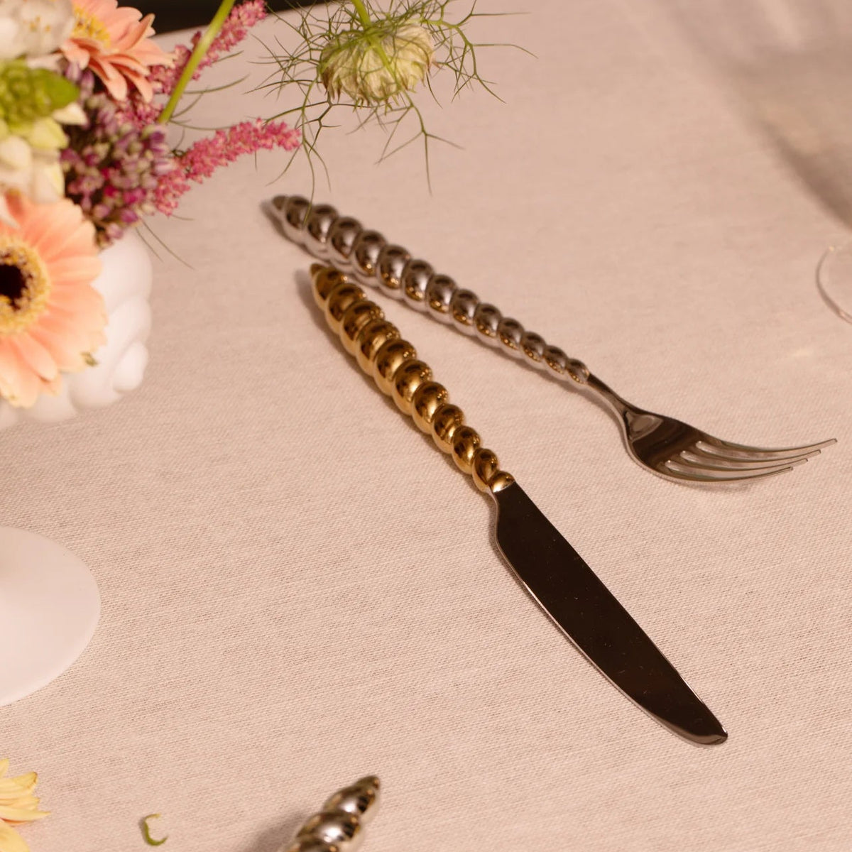 A gold-tone dinner knife and a silver-tone fork from a L’Objet Jessica McCormack Shell Flatware 5 pc Set, featuring textured, shell-like handles resting on a light-colored tablecloth near a small vase of flowers.