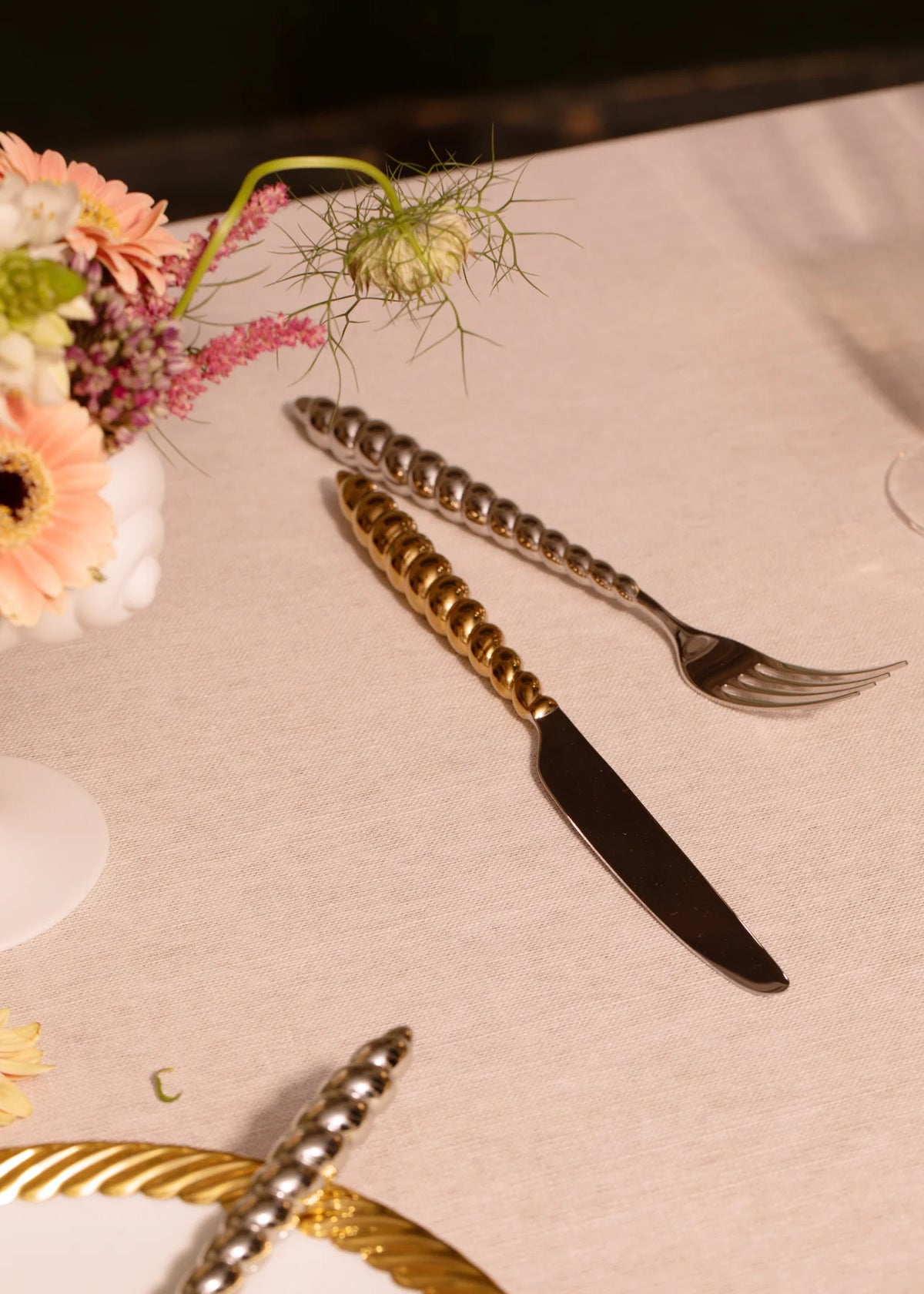 A gold-toned knife and silver-toned fork from a L’Objet Jessica McCormack Shell Flatware 5 pc Set - Gold, both with spiral-textured handles, rest on a linen tablecloth next to a small floral arrangement.