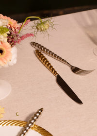 A gold-toned knife and silver-toned fork from a L’Objet Jessica McCormack Shell Flatware 5 pc Set - Gold, both with spiral-textured handles, rest on a linen tablecloth next to a small floral arrangement.