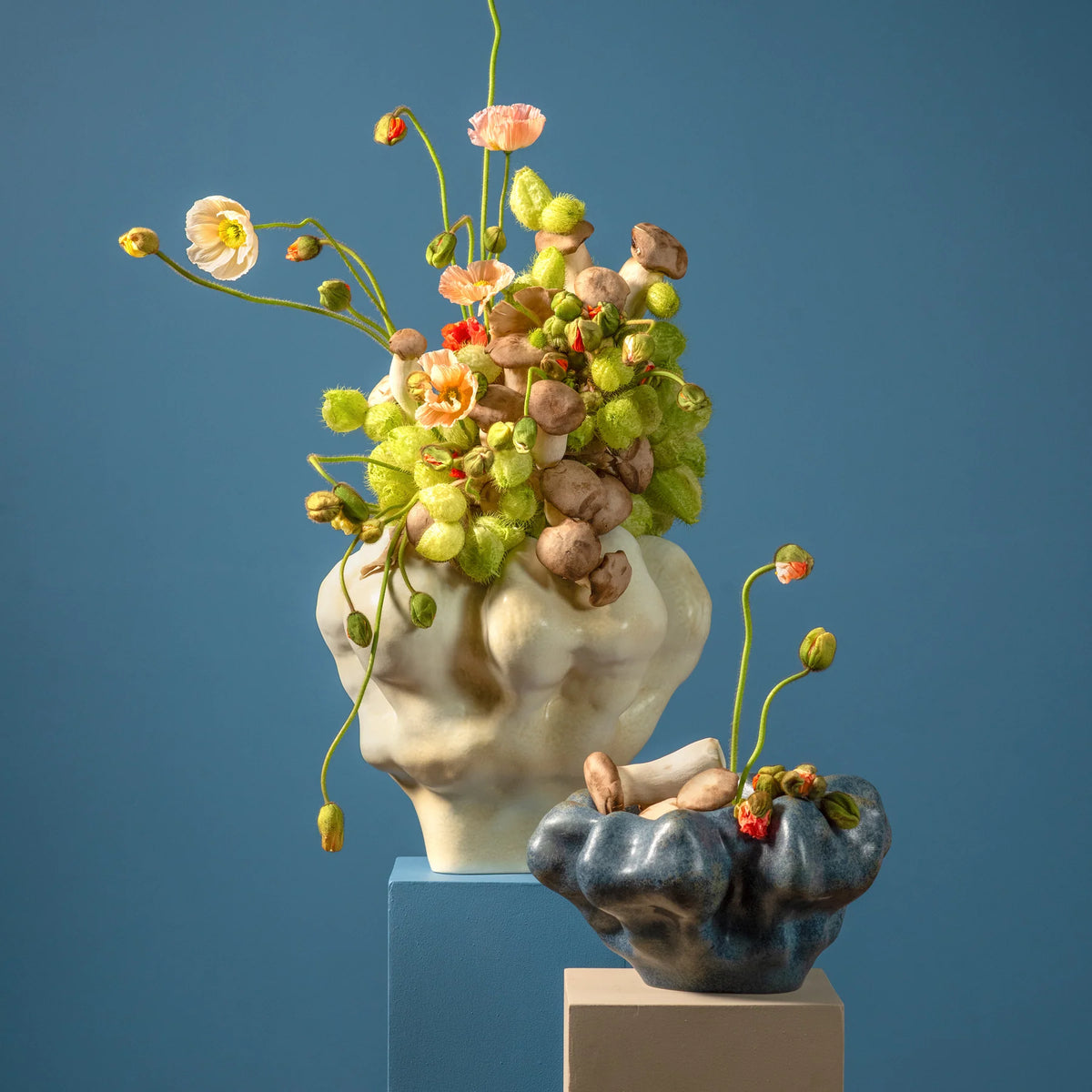 Two Timna Coupe vases by L’Objet, crafted in an aged iron finish and featuring wavy, organic forms, sit atop pastel blocks. Their stunning reactive glaze catches the light—the taller vase displays poppies and seed pods, while the smaller one holds just a scattering of pods. The scene is set against a solid blue background.