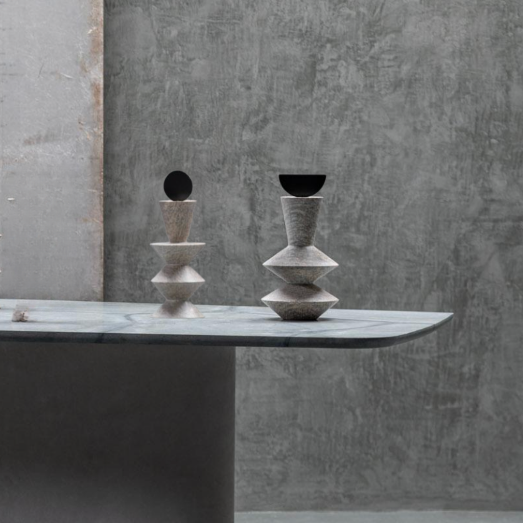 Two Constantin Gray w/ Black Sculptures by Gardeco, featuring geometric forms and dark circular tops, sit on a marble-topped table against a gray textured concrete wall, embodying the essence of contemporary home decor and modernist sculpture.