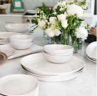 Relish’s Weave Melamine Dinnerware in white is neatly stacked on a marble countertop, with a bouquet of white roses and greenery in glass vases nearby as someone writes in a notebook.