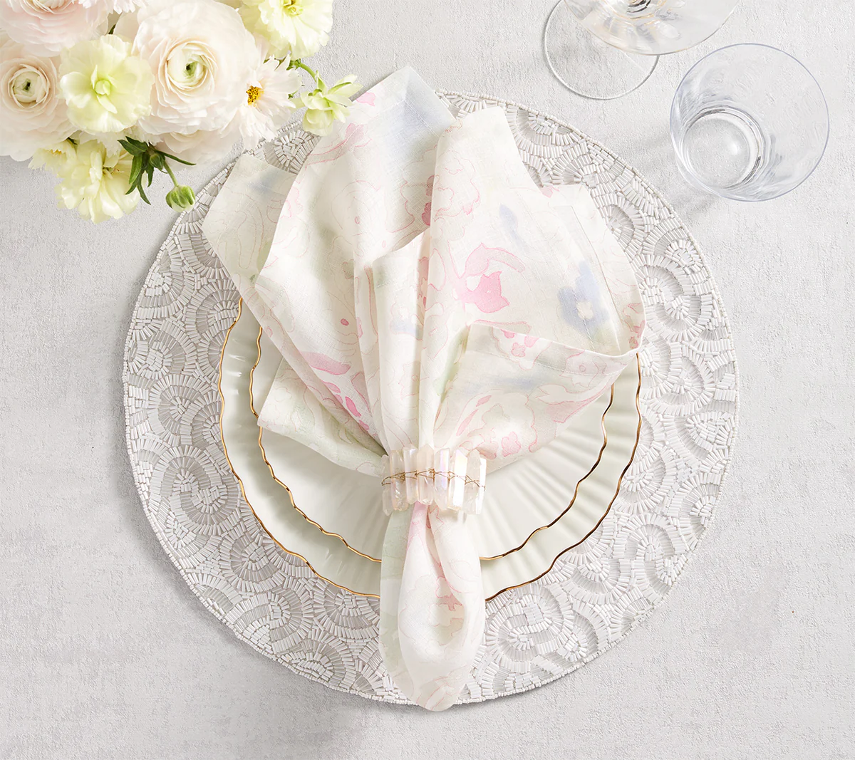 Elegant table setting with a white embossed charger, scalloped white plate with gold trim, Kim Seybert Giverny Napkin Mint and Multi in a ring, clear glasses, and a bouquet of white flowers.