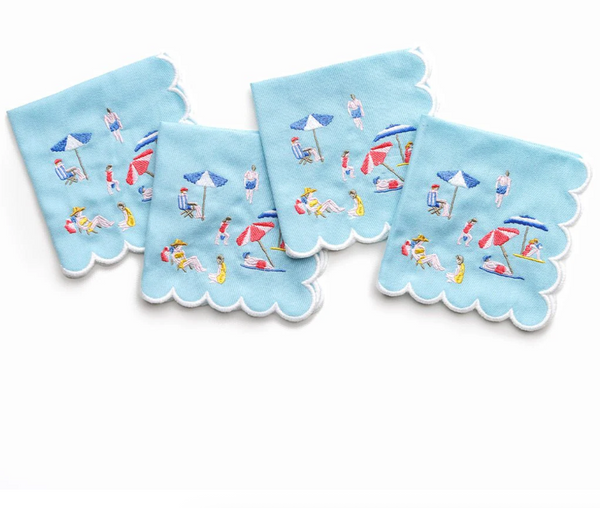 Day At The Beach Cocktail Napkin s/4 by Pomegranate features four light blue scalloped-edge napkins embroidered with beach scenes—people, umbrellas, and towels—perfect for beach-themed tableware or colorful decor. Displayed overlapping on a white background.