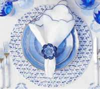 Elegant table setting with blue and white patterned plates, a white napkin with navy trim in the Kim Seybert Pareo Napkin Ring Set of 4 - Periwinkle & Blue, white-handled cutlery, blue floral glassware, and an embroidered tablecloth.
