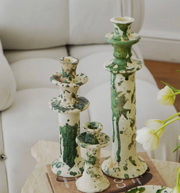 Three Vintage Splatter Candlestick Green by Moroccan White stand on a beige table beside a brown book and white tulips, creating an artistic table setting with a white couch in the background.