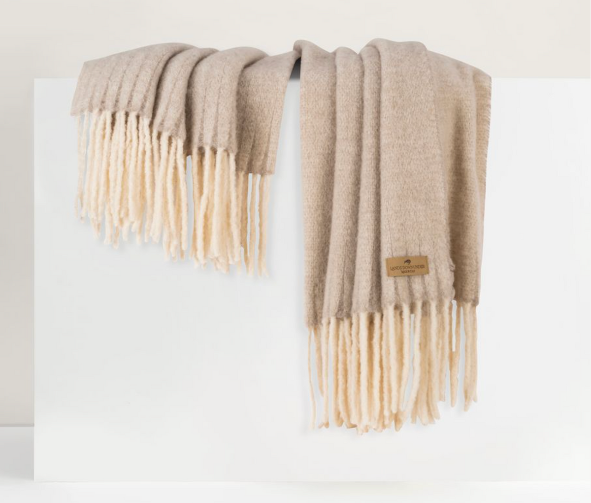 The Andes Throw by Lands Downunder, a soft beige alpaca wool blanket with thick cream fringe and a small brown tag, is draped over a white rectangular surface against a minimal light-colored background.