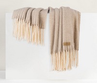 The Andes Throw by Lands Downunder, a soft beige alpaca wool blanket with thick cream fringe and a small brown tag, is draped over a white rectangular surface against a minimal light-colored background.
