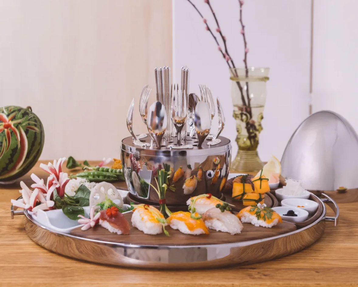 A round MOOD PARTY Christofle Stainless Steel Round Tray 15.7″ (40cm) holding a central set of upright cutlery and a wooden board arranged with various pieces of sushi.