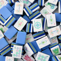 A vibrant assortment of Mahjong tiles from the Oh My Mahjong brand’s Moonlight Tiles Set, primarily in blue and white hues and adorned with intricate illustrated symbols, numbers, and floral motifs. Crafted from high-quality acrylic, these stylish tiles are artfully scattered and overlapping—an exceptional choice as unique game accessories for any collection.