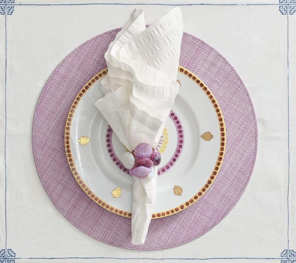 A white napkin in a Kim Seybert Sea Stone Napkin Ring Set of 4 - Lilac rests on a white plate with gold accents, atop a lavender charger and a white tablecloth with light blue embroidered corners.