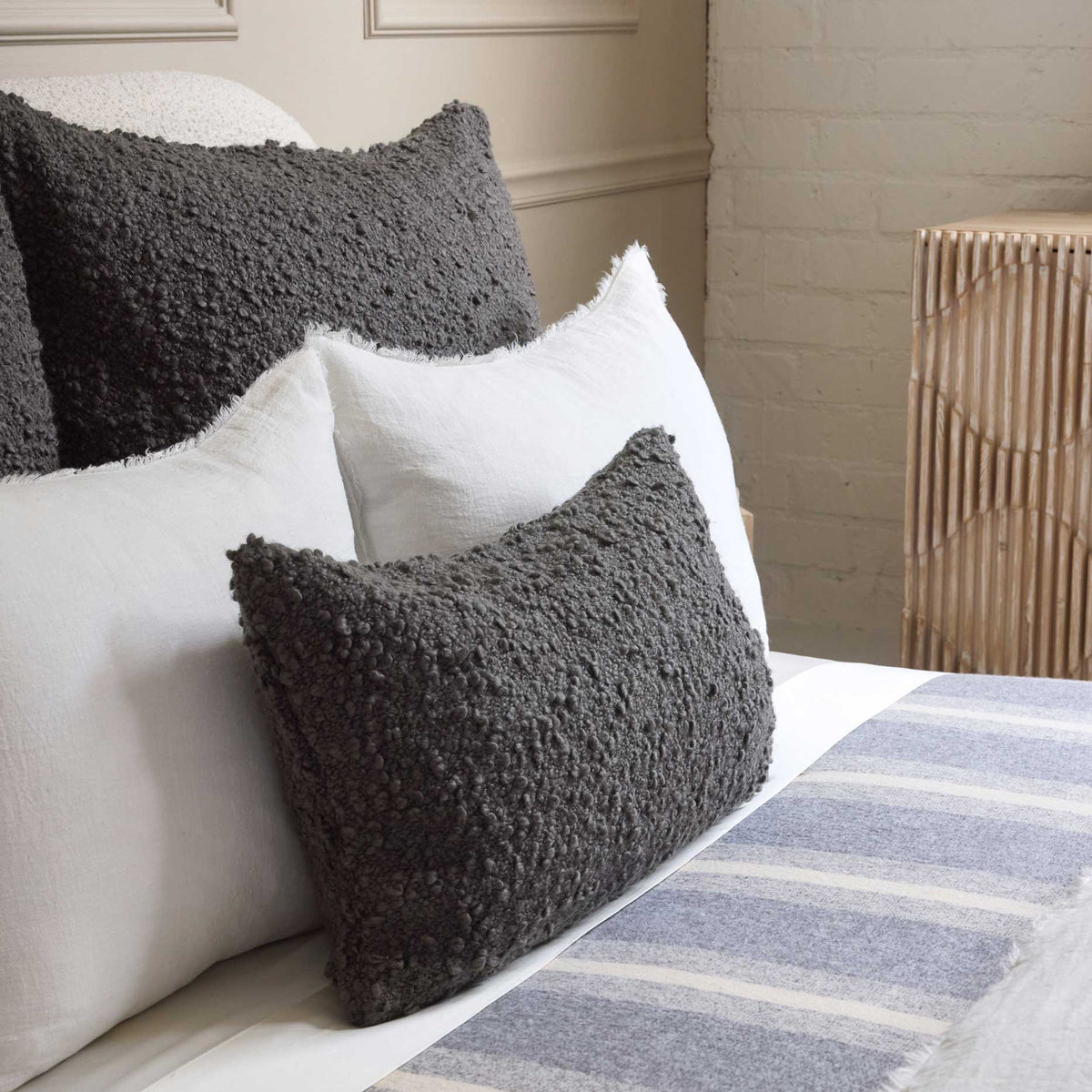 A neatly made bed with Pom Pom At Home's Murphy Pillow Midnight 14 x 24, a striped gray and white blanket, and a wooden nightstand against a cream wall creates an inviting, cozy home atmosphere.
