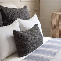 A neatly made bed with Pom Pom At Home's Murphy Pillow Midnight 14 x 24, a striped gray and white blanket, and a wooden nightstand against a cream wall creates an inviting, cozy home atmosphere.