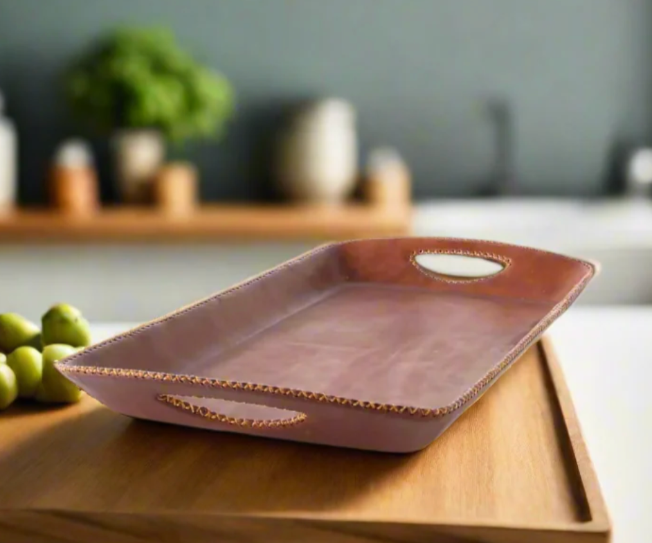 The Embroided Long Bar Tray - Brown by Bati Goods, featuring gold-trimmed edges and handles, sits atop a wooden surface. Crafted from organic tanned leather for a luxurious touch, its water-resistant base is adorned with green grapes, while a blurred kitchen background completes the scene.