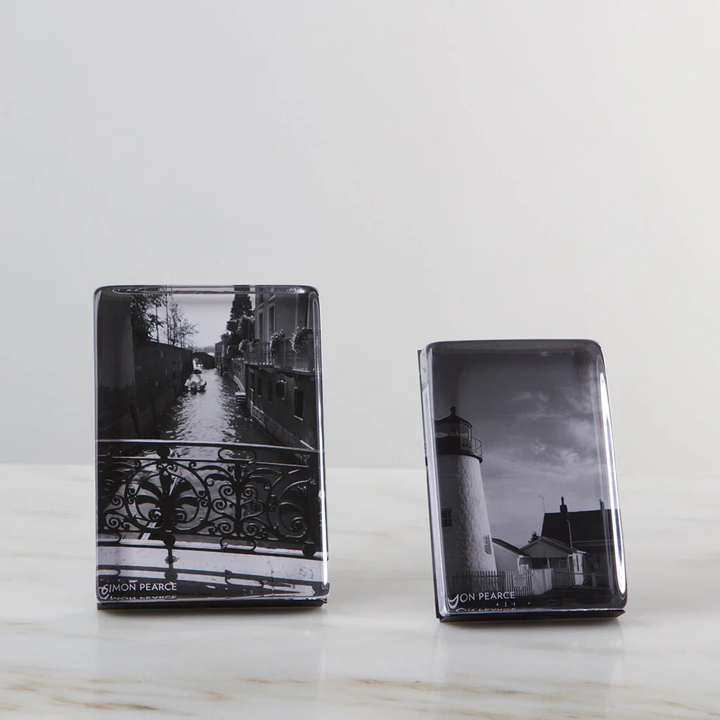 Two Simon Pearce Woodbury Photo Block Verticals sit on a marble surface. The left holds a black-and-white canal scene with a bridge, while the right displays a lighthouse and house, both in dramatic monochrome.