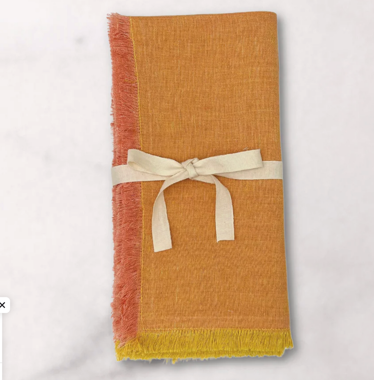 The Lia Napkin Set of 4 - Pomelo by Be Home features folded orange yellow napkins with frayed edges, pink and yellow fringe, and a cream ribbon bow, adding charm to your table setting against a light background.