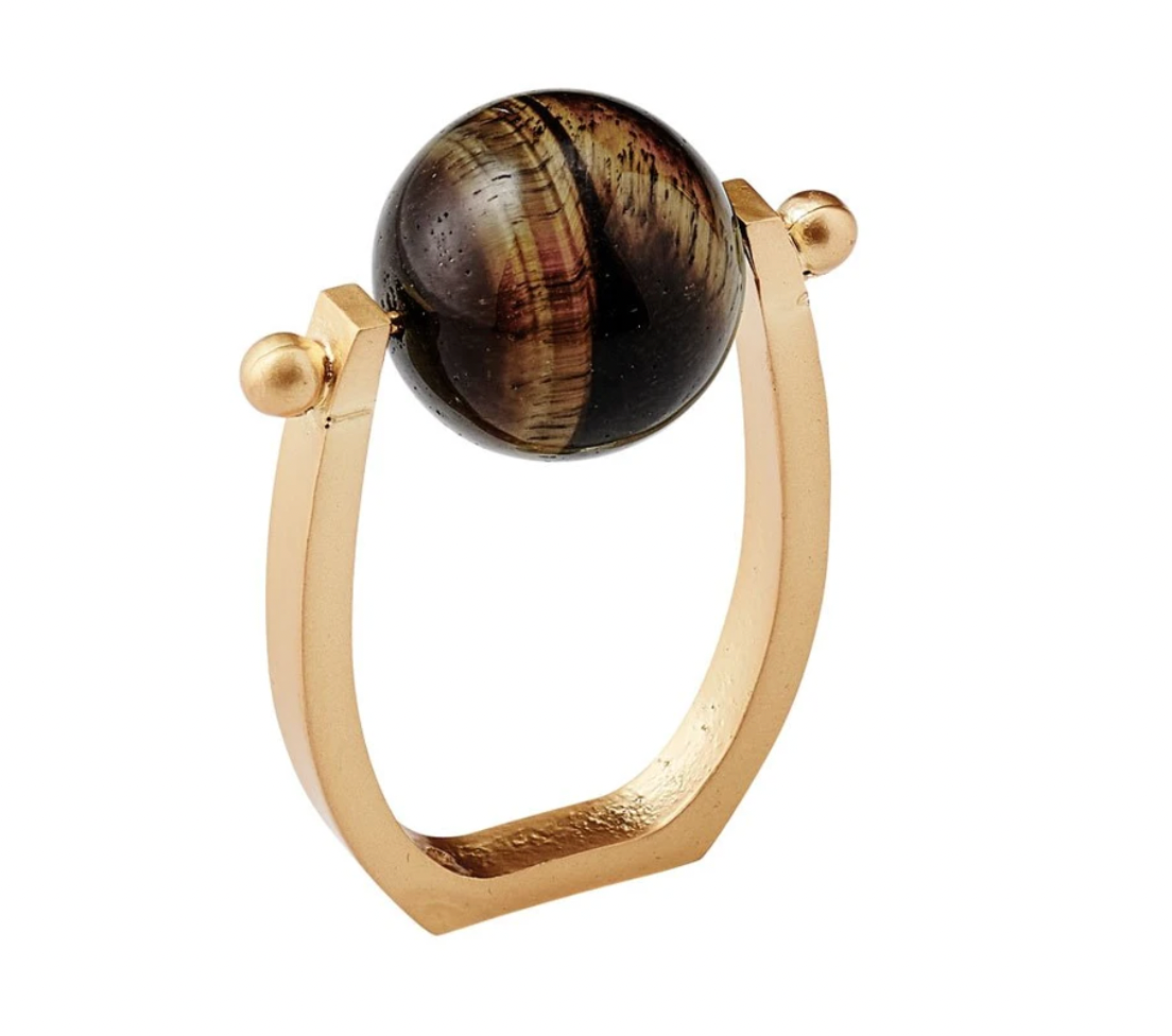 The Mineral Napkin Ring s/4 - Brown by Kim Seybert showcases a sophisticated design with a rectangular open gold band and a striking, large round dark tiger eye stone set between two small gold spheres on top. This elegant napkin ring lends a refined and stylish accent to any table setting.