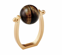 The Mineral Napkin Ring s/4 - Brown by Kim Seybert showcases a sophisticated design with a rectangular open gold band and a striking, large round dark tiger eye stone set between two small gold spheres on top. This elegant napkin ring lends a refined and stylish accent to any table setting.