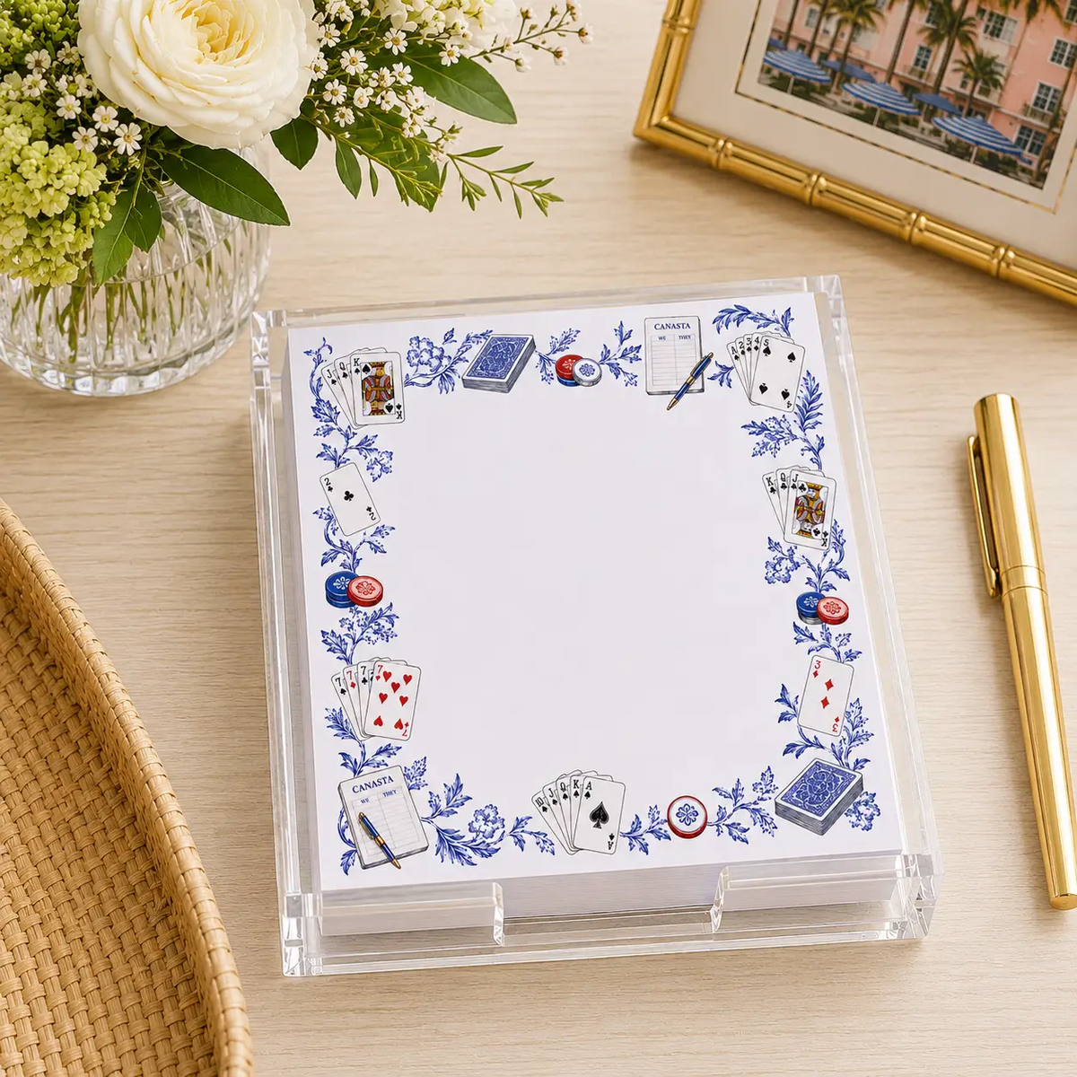 A clear acrylic holder containing a stack of paper for the Maven Home Proper Canasta Notepad & Holder, decorated with a blue floral border featuring illustrated playing cards, scorecards, and poker chips.