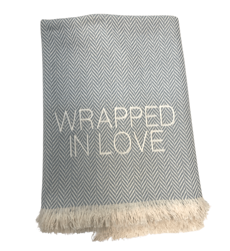 The Wrapped in Love Throw by A Soft Idea features a grey and white herringbone pattern, beige fringe, “WRAPPED IN LOVE” embroidery, is super soft, machine washable, and makes a perfect cozy holiday gift.