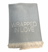 The Wrapped in Love Throw by A Soft Idea features a grey and white herringbone pattern, beige fringe, “WRAPPED IN LOVE” embroidery, is super soft, machine washable, and makes a perfect cozy holiday gift.