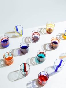 Fifteen Gesti Tumblers by Ichendorf Milano, each filled with colorful liquids, are arranged in rows on a white surface, casting long, multicolored shadows.